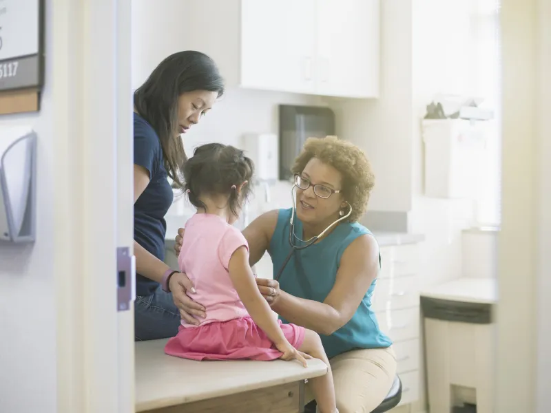 A female AHS doctor listens to a little girl's heart in the exam room while her mother comforts her