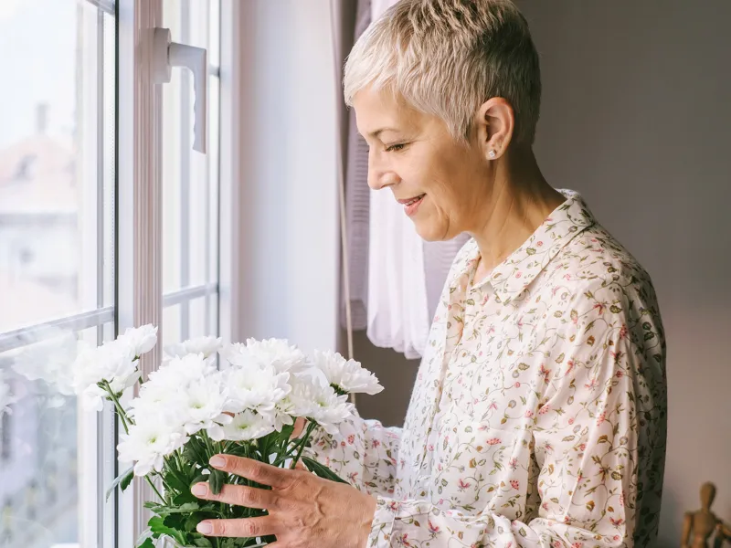 An adult caucasian woman arranges a vase of flowers by the window.