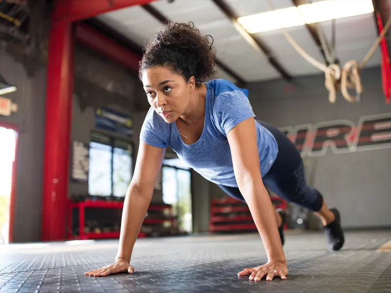 A young African American woman with a blue shirt performs push-ups at an open spaced gym.