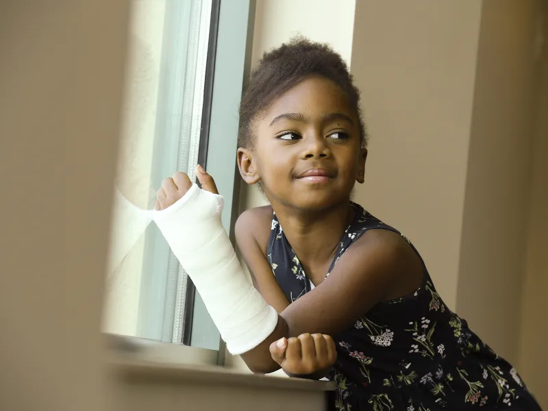 A little girl in a cast smiles while sitting next to a windowsill