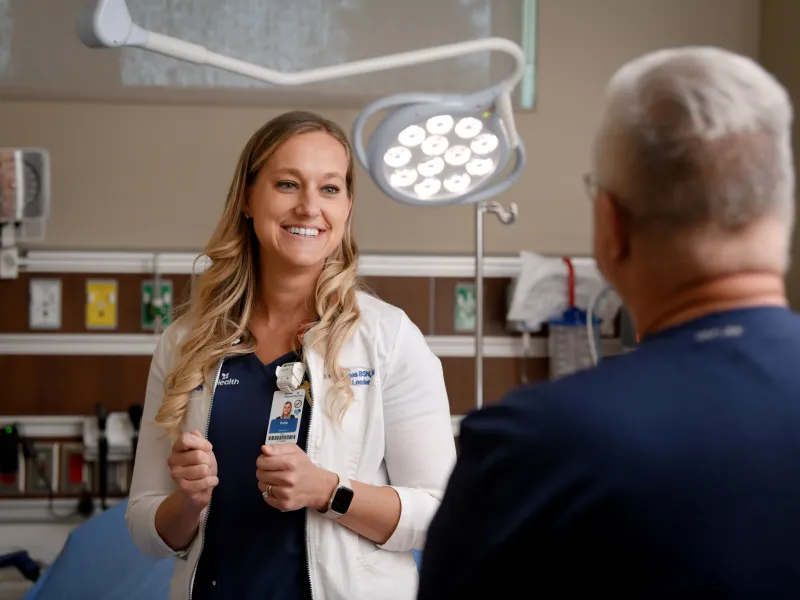 A healthcare professional in a white coat is speaking with a patient in a hospital room. Medical equipment and a bed are visible in the background.