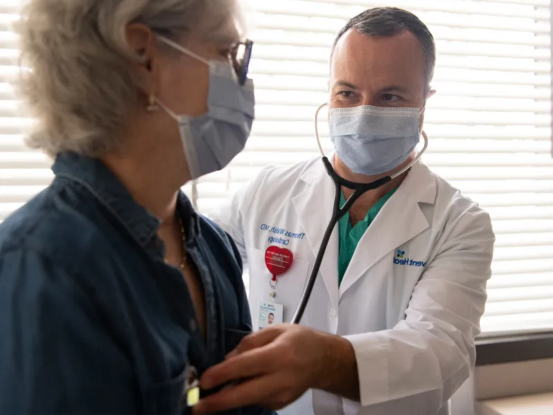 Doctor listening to patient's heartbeat with a stethoscope while both wear masks.