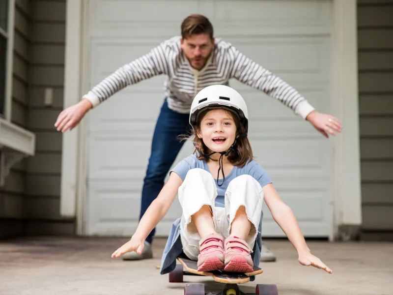 A dad showing his little girl how to skateboard in the carport.