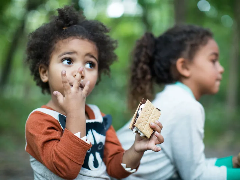 Kids eating s'mores while seating around the campfire in the forest.