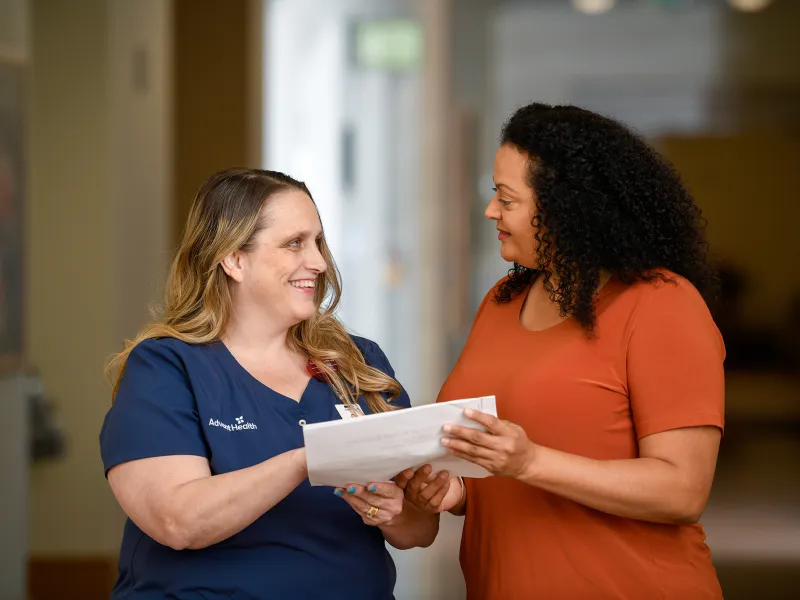 An AdventHealth nurse talking with a female patient while both hold a document.