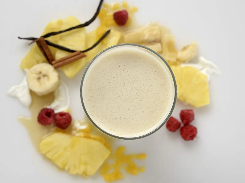 a creamy smoothie in a glass, surrounded by slices of pineapple and banana with vanilla bean pods