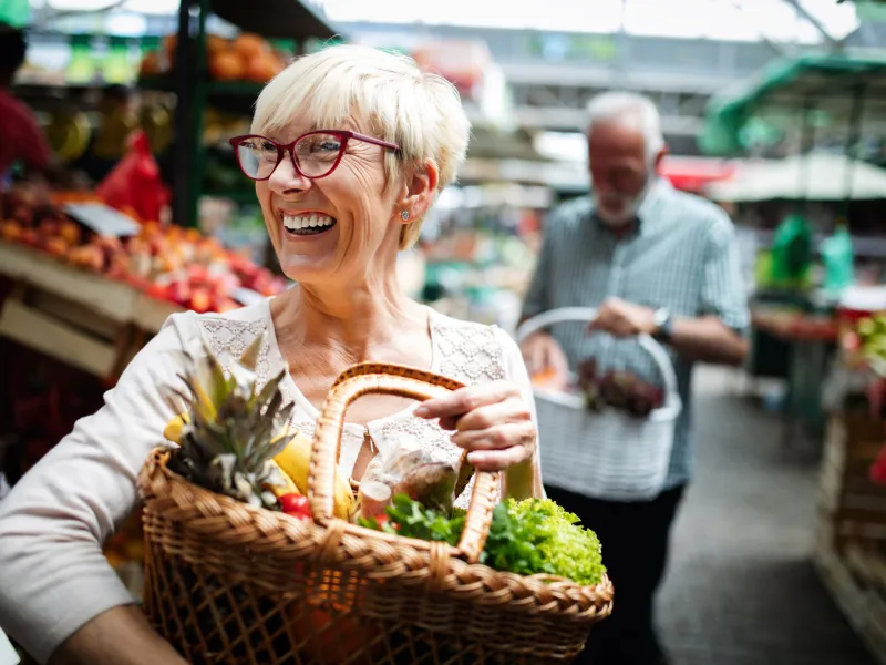 Woman smiling in the foreground while her husband is in the background. Both are holding a basket of fresh produce at a farmers market.