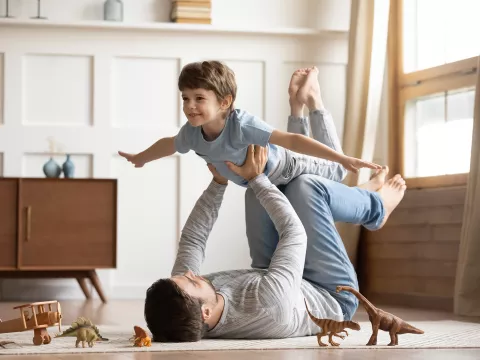 A Little Boy Plays "Air Play" on his dad's legs on the floor with dinosaur toys surrounding them