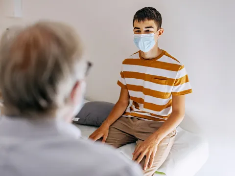 A Teenager Wearing a Face Mask Speaks to His Physician.