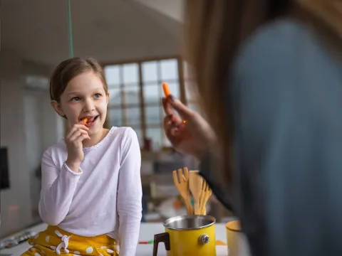 A Mother Hands Her Daughter Carrot Sticks to Eat in the Kitchen.