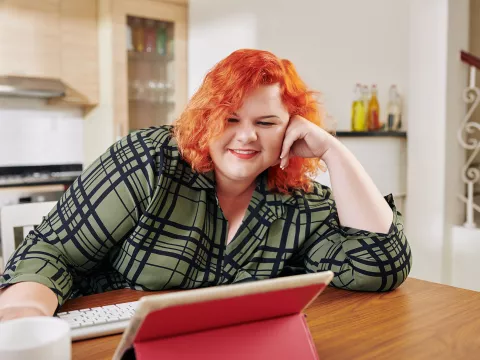 A Woman Smiles as She Sits at Her Kitchen Table Viewing Content on Her Tablet.