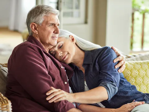 A Senior Woman Rests Her Head on Her Senior Husbands Shoulder While They Sit On a Couch at Home