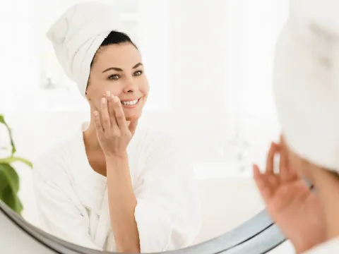Woman in white bathrobe and towel smiling in mirror while applying skincare cream to her face in a bright bathroom