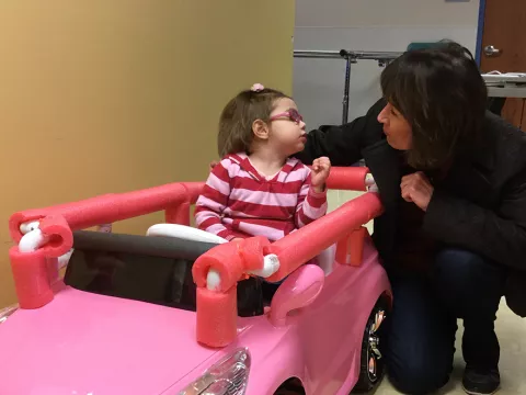 A little girl simulating to drive a pink car toy while kissing her mother.