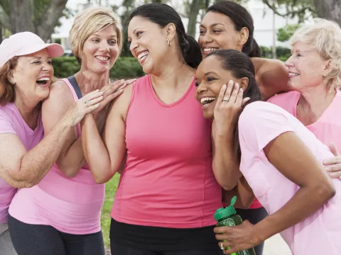 A group of friends, all wearing pink and hugging a woman in the venter, to support a breast cancer survivor.