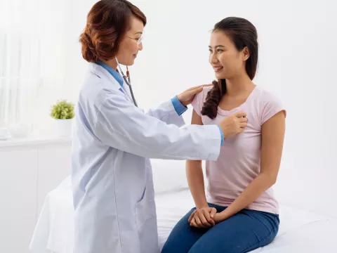 A woman is sitting on an exam table while a doctor listens to her heart.