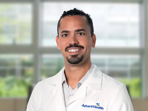 Dr. Paoli, a tan-skinned man with dark hair and facial hair, smiles at the camera. Dr. Paoli is wearing a white medical coat, and he stands in front of a blurred, office-style background.