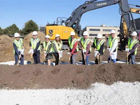Dignitaries wearing hard hats use shovels to dig into pile of dirt in front of construction equipment during groundbreaking ceremony.
