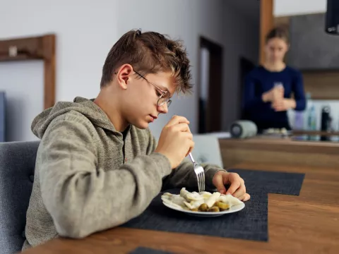 A boy sits at a dining room table eating lunch in the foreground while his mother is in the background adding seasoning to food on a plate.