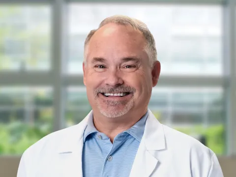 Chris Nelson, DC, smiles at the camera in front of a blurred office-style background. He is a white male with short grey hair and light facial hair. He is wearing a white medical coat and a blue button-up shirt. 