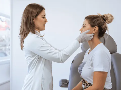 Dermatologist examining female patient's face in room