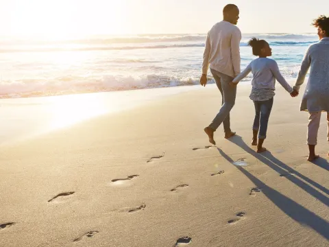 A family of three walks along the beach at sunset leaving footprints in the sand.