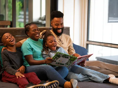 A family laughs while reading a book together as they are seated on the floor.