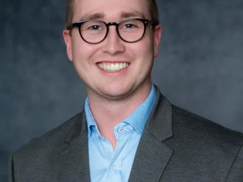 Hudson Sutton smiles at the camera in front of a grey backdrop. He is a white man with red hair. He is wearing brown-rimmed glasses, a blue dress shirt, and a grey sports coat.