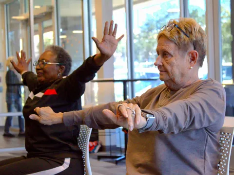 Two seated senior women stretch out their arms in a classroom during a wellness class.