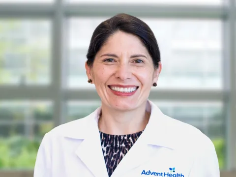 Dr. Elise Jaramillo-Mayor smiles at the camera in front of a blurred office background. She is a pale-skinned woman with dark hair, and she is wearing an animal print shirt and a white medical coat. 