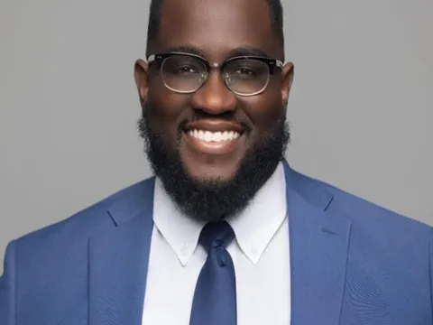 Mack Vendome, an African-American man wearing a blue suit, white shirt, and glasses, smiles in front of a plain gray background.