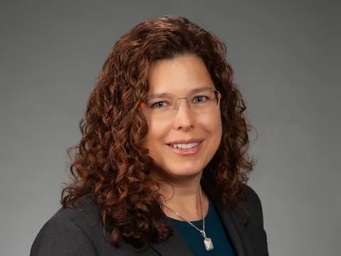 Marcy Cent, the new director of oncology services for AdventHealth Georgia, smiles at the camera in front of a grey backdrop. Marcy is a white woman with curly auburn hair. She is wearing glasses.