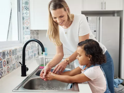 Mother showing her daughter how to wash her hands at the kitchen sink.