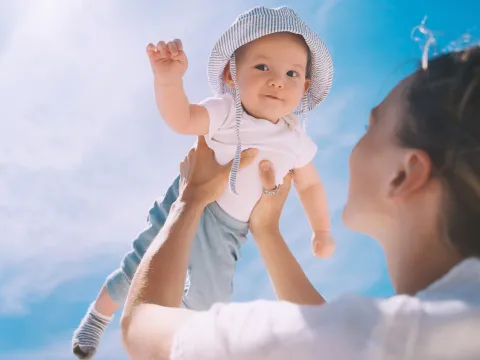 A mother holds up her baby whose wearing a hat while outside.