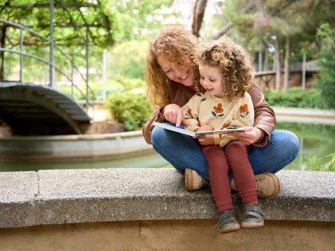 Mother and child reading a book together while sitting on a wall by a river.