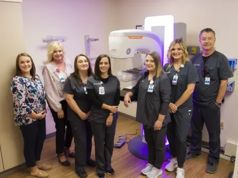 The AdventHealth Murray breast imaging team is pictured in the mammography room. There is a 3D mammography machine behind a group of people in gray scrubs. Two of the people are in professional wear.