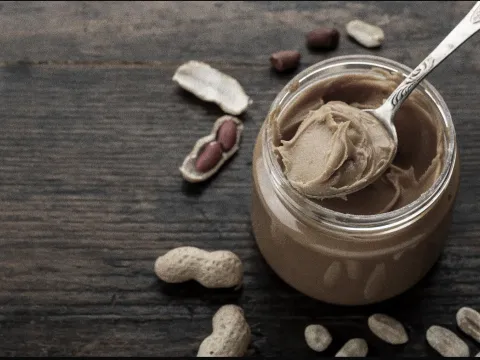 A spoonful of peanut butter sitting on a jar of peanut butter and peanuts surrounding the jar on a table.