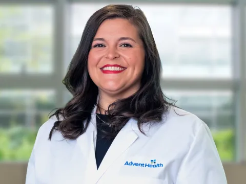 Alison Petty, NP-C, smiles at the camera in front of a office-style background. She is a white woman with brunette hair wearing a black shirt and a white medical coat. 