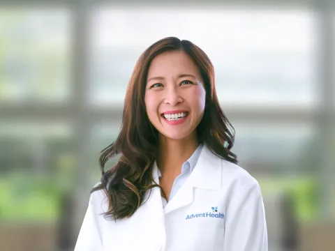 Phoebe Carpenter, an Asian woman with dark hair, smiles at the camera. She is wearing a white medical coat, and she stands in front of a blurred, office-style background.