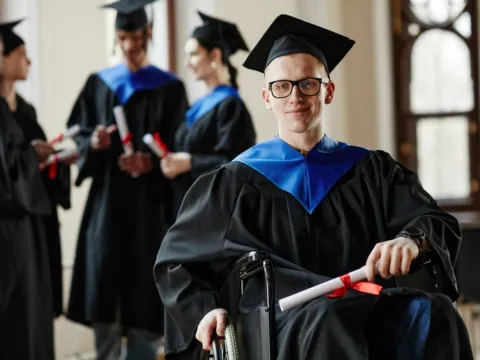 A student in a wheelchair wears a graduation cap and gown and holds a diploma.