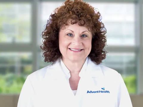 Valorie Mixon, a physician assistant, smiles at the camera in front of a blurred office-style background. She is a white woman with curly brown hair and blue eyes. She is wearing a white AdventHealth medical coat.