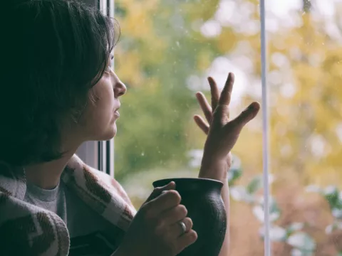 Woman looking out and touching window while indoors while wrapped in a blanket and holding a mug.