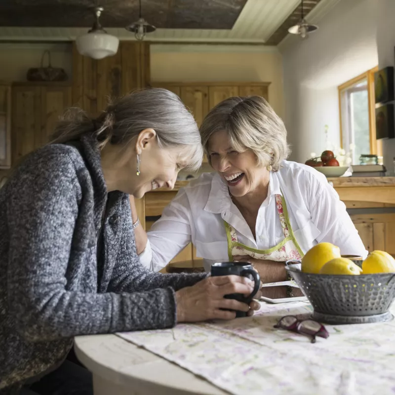 Two adult Caucasian women laugh together at a dining room table.