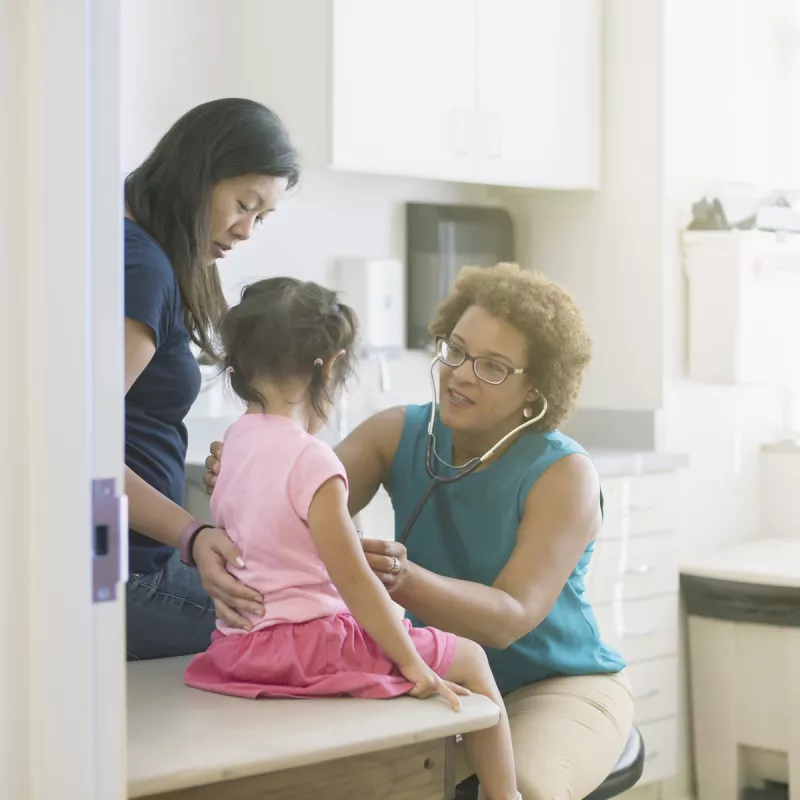 A female AHS doctor listens to a little girl's heart in the exam room while her mother comforts her
