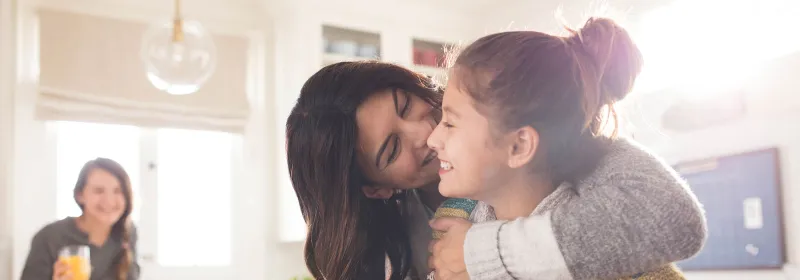 A family of three women are in the kitchen, one daughter hugging the youngest while she flips pancakes.