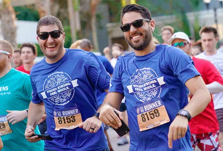 Two men smile at the camera while running Track Shack's annual Corporate 5k. Their t-shirts say, "Finish Strong, Feel Whole."
