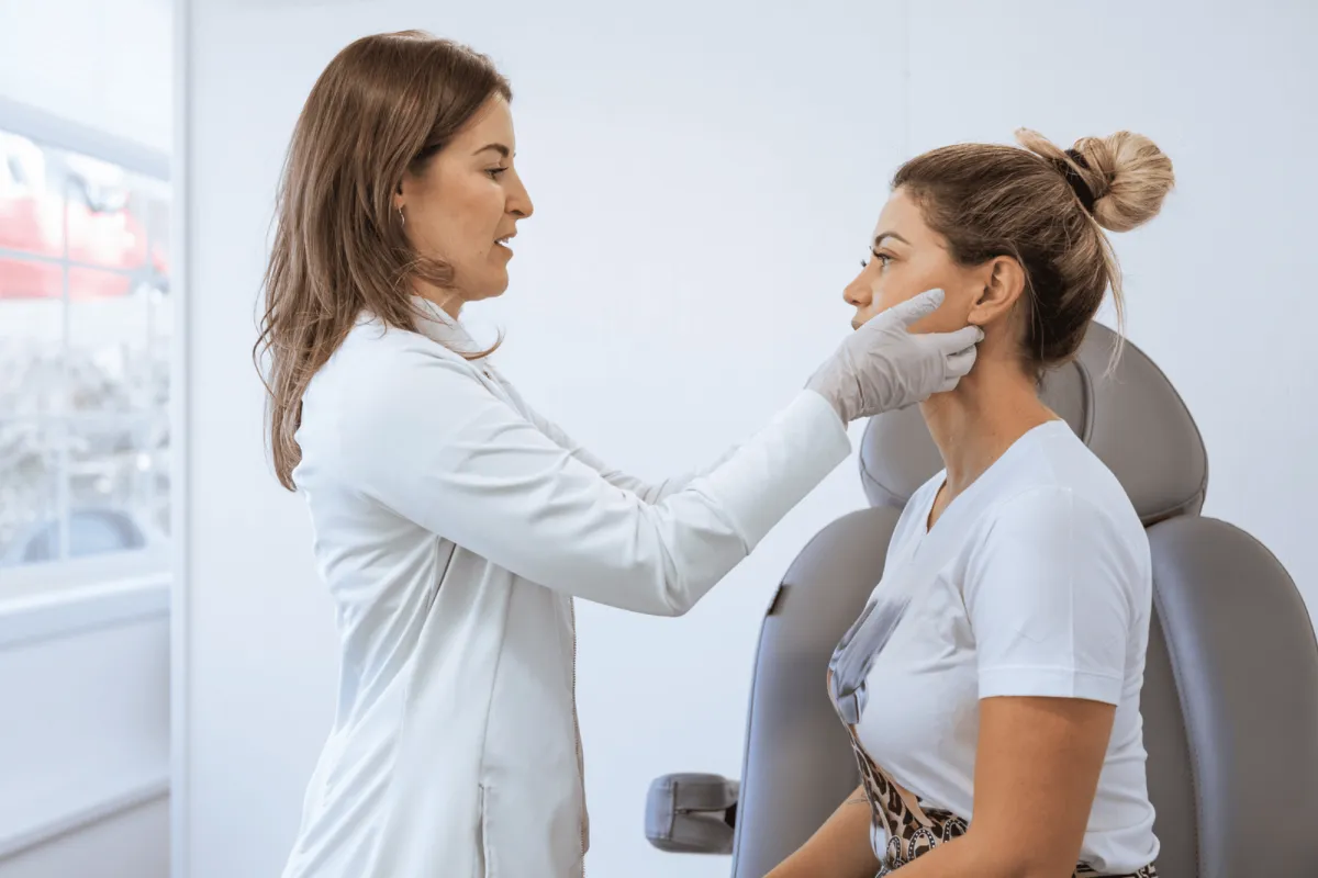 Dermatologist examining female patient's face in room