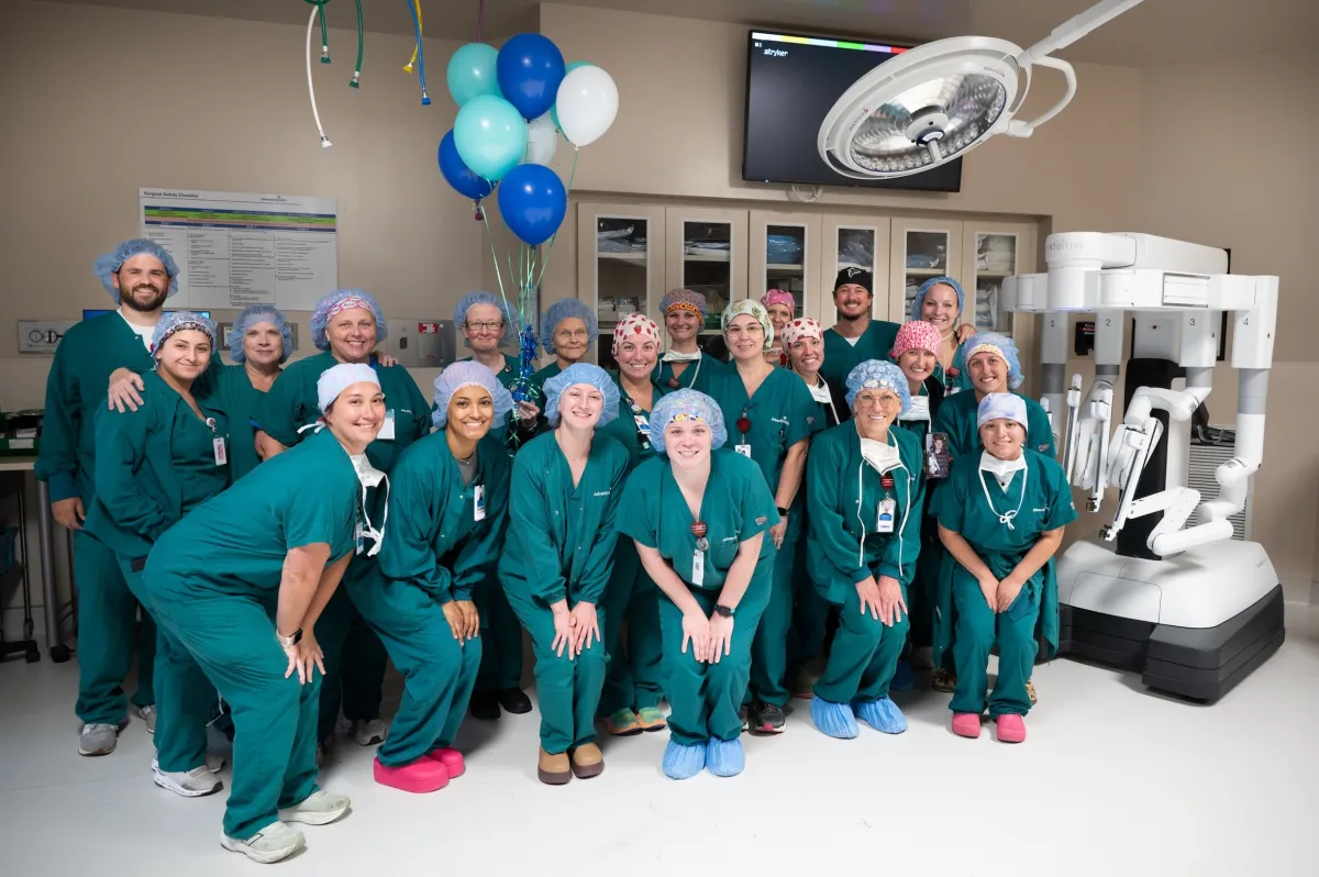 A large group of people in teal scrubs stand in a surgical suite with a white surgical robot. There is a small cluster of balloons in different shades of blue behind them.