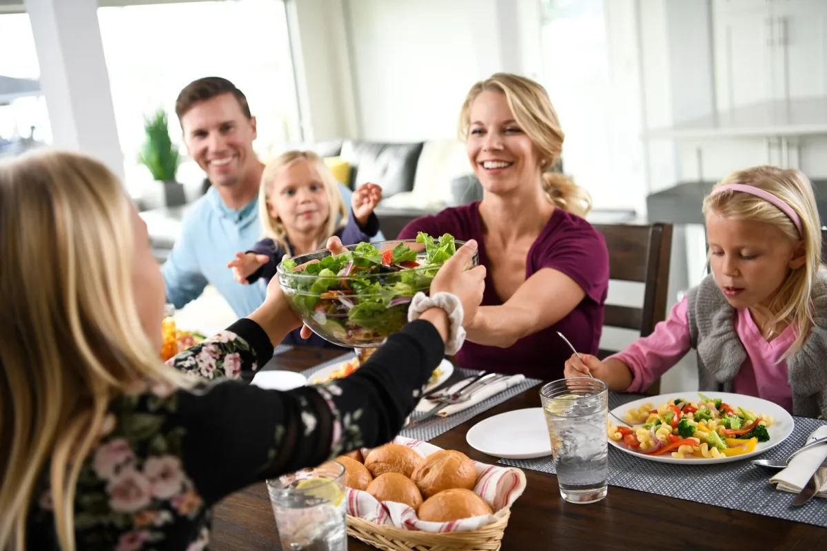 A woman hands a salad to a girl as a family shares a meal at the dinner table.