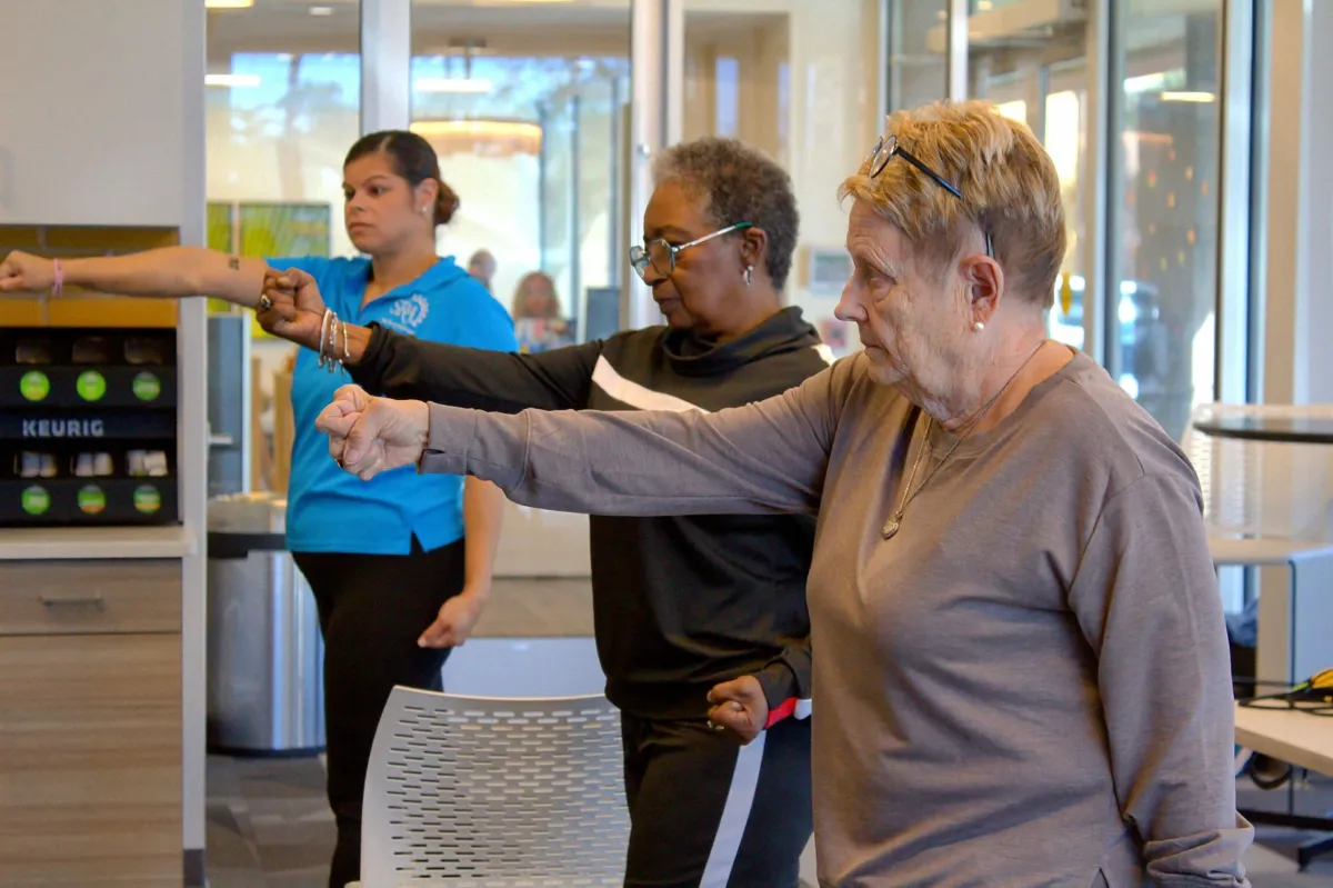 Two senior women participate in a balance class, with a female instructor in the background.